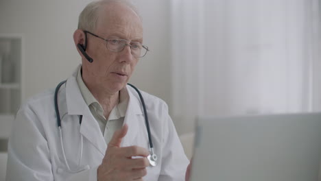 Der-ältere-Facharzt-Kommuniziert-Per-Webcam-Auf-Dem-Laptop-Von-Seinem-Büro-In-Der-Klinik-Aus-Und-Kommuniziert-Aus-Der-Ferne-Mit-Patienten